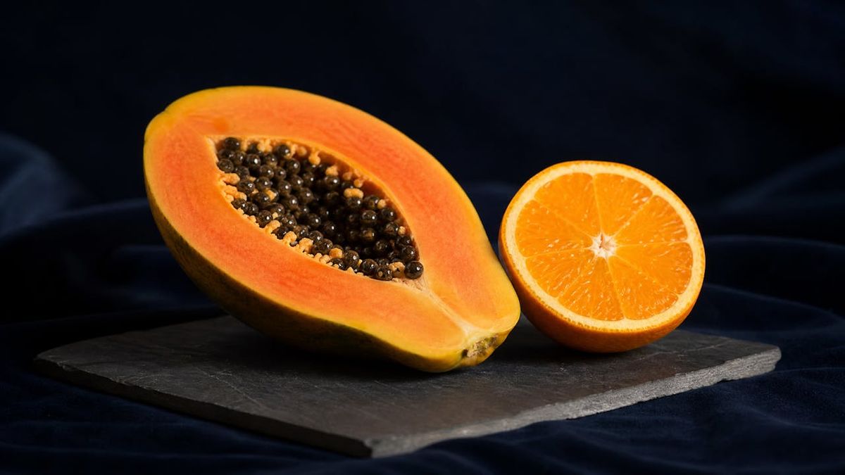 A halved papaya and a halved orange on a dark slate board