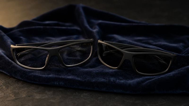 Two pairs of black wraparound UV-protective glasses, one clear and one tinted, on navy velvet
